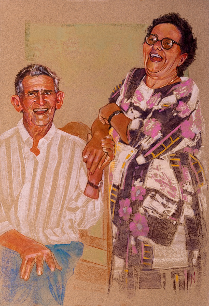Retrato de corpo inteiro de um casal idoso rindo, ele sentado, ela em pé de vestido florido.